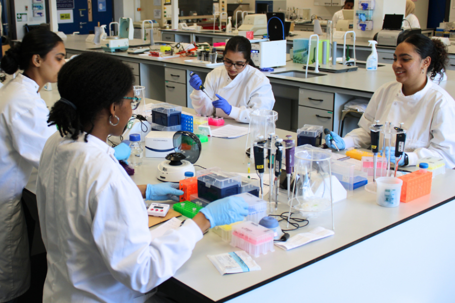 4 female students wearing lab coats are in a laboratory sat around a bench. There is an array of lab equipment on the bench and some students are using the equipment.
