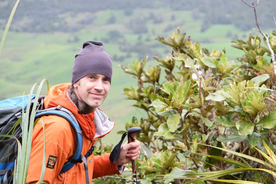 Robbie hiking, smiling standing next to a woody shrub