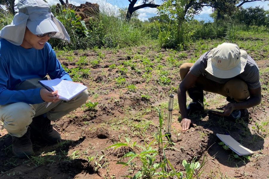 Two students kneeling in the soil with tools and plants, taking notes in a notebook