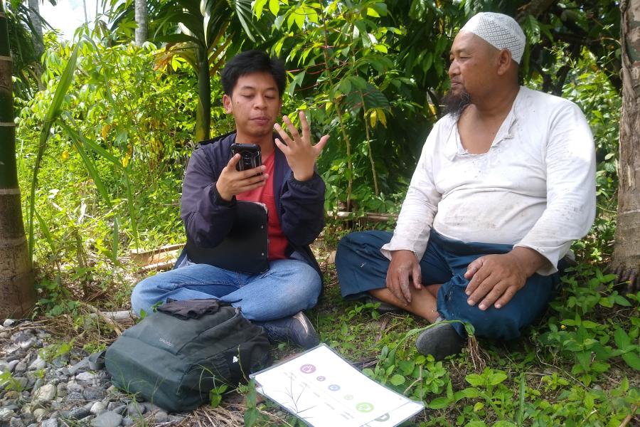 Two Indonesian men sat cross legged in the shade with foliage in the background. They are discussing a document that is in front of them. The older one is happily watching the younger one gesticulate with his hands 