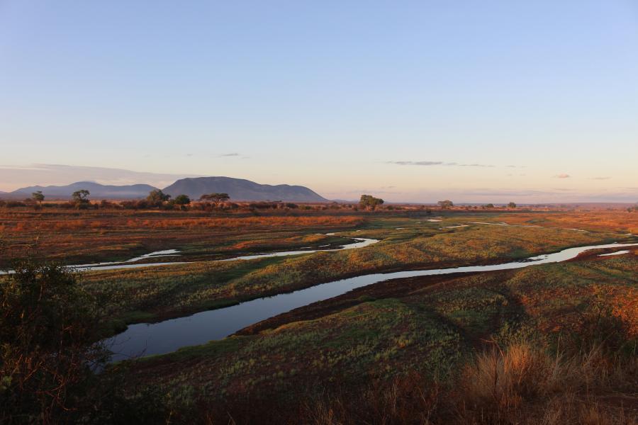 photo of a landscape in Tanzania with a slow river in the foreground and a mountain in the distance. The image is backlight by low orange sunlight 