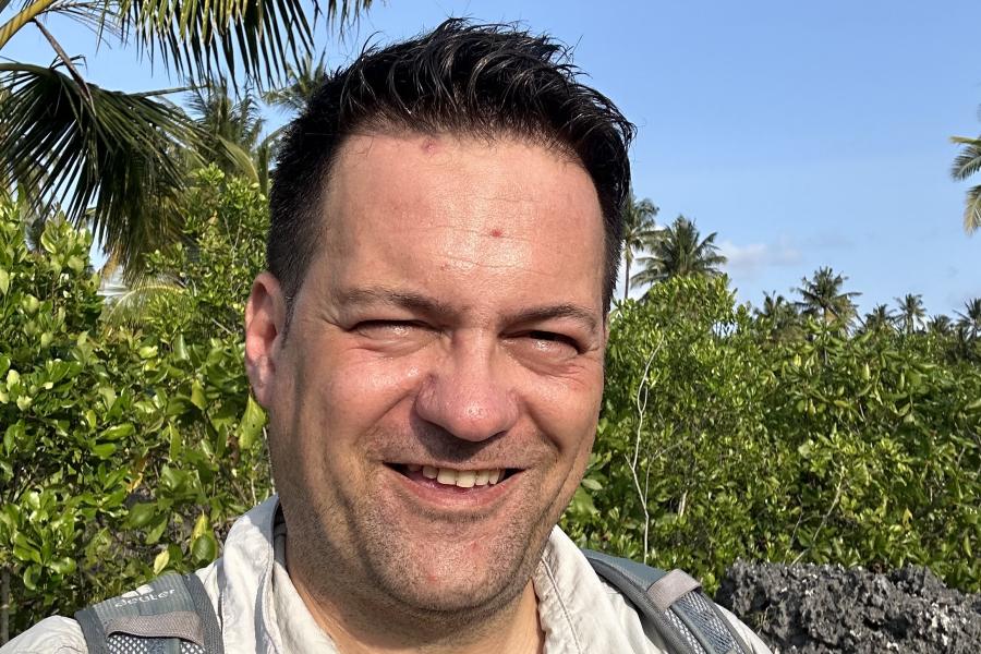 Alex standing with palm trees and vegetation in the background