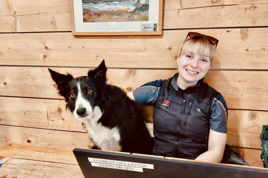 Jessica sitting in front of a wooden cabin wall with a sheepdog.