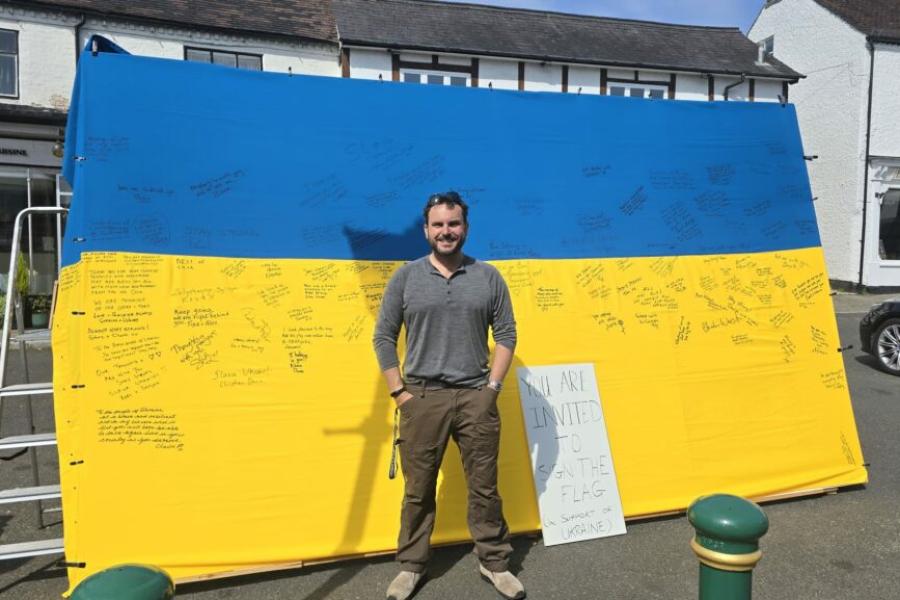 A person stands in front of a large blue and yellow flag, resembling the Ukrainian flag, covered with handwritten messages. A nearby sign reads, 'You are invited to sign the flag. Support Ukraine.'