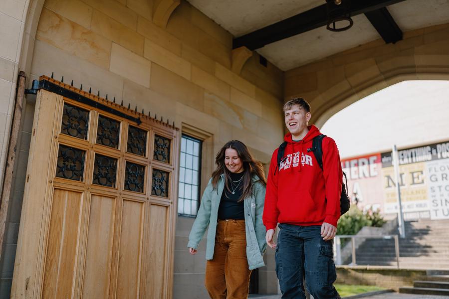 Two students walking through memorial arch one wearing a red Bangor University hoodie the other a blue coat