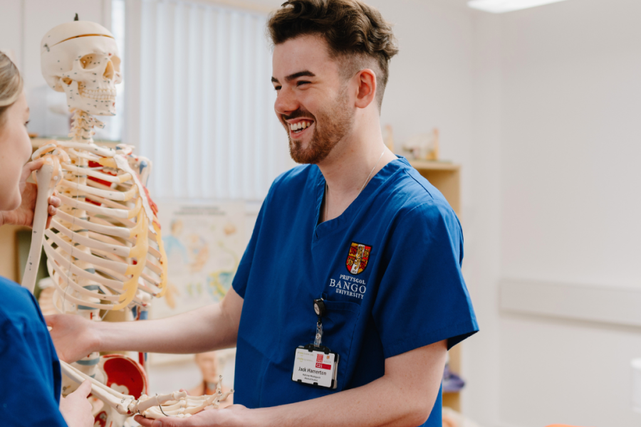 a male student doctor wearing blue scrubs looking at an anatomical model of a skeleton