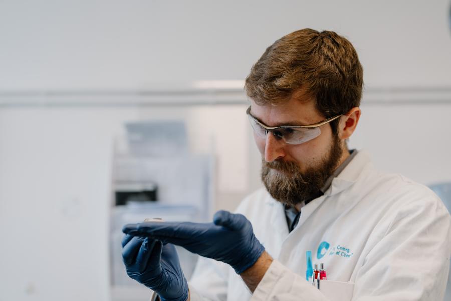A man in a lab wearing glasses and gloves looking at a object in his hand