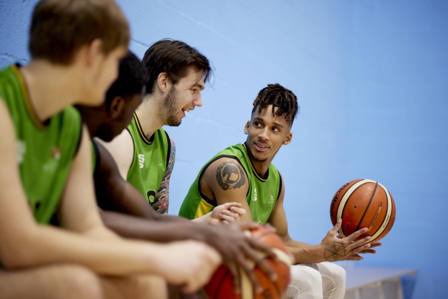 Myfyrwyr yn eistedd ar y meinciau yn sgwrsio yn un o'r neuaddau chwaraeon yng Nghanolfan Brailsford. Mae un yn gafael mewn pêl fasged.