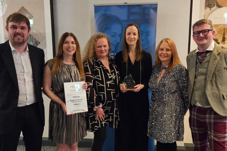 A group of six people standing indoors in front of a banner, dressed in formal and semi-formal attire. Two individuals in the centre are holding awards: one holds a framed certificate that reads “HELOA Awards Winner – Regional Outreach and Recruitment Award,” and the other holds a glass trophy. The background includes framed artwork and a blue banner.