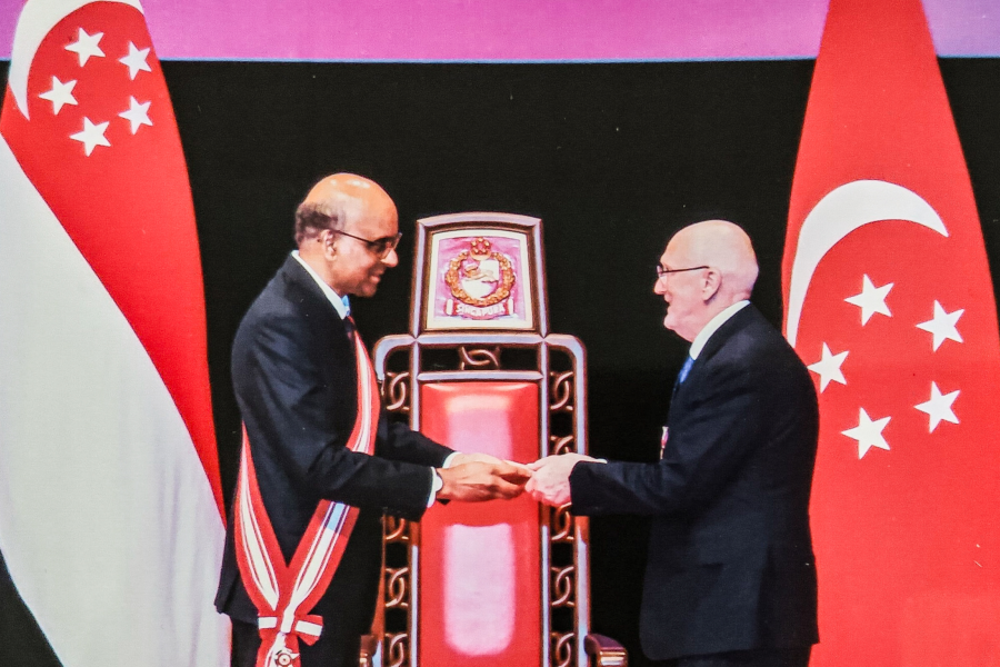 Two individuals stand on a stage in front of a large ornate chair with the Singapore coat of arms above it. They are shaking hands while one presents an object to the other. Both are dressed formally, and one wears a ceremonial sash. Two large Singapore flags flank the scene.