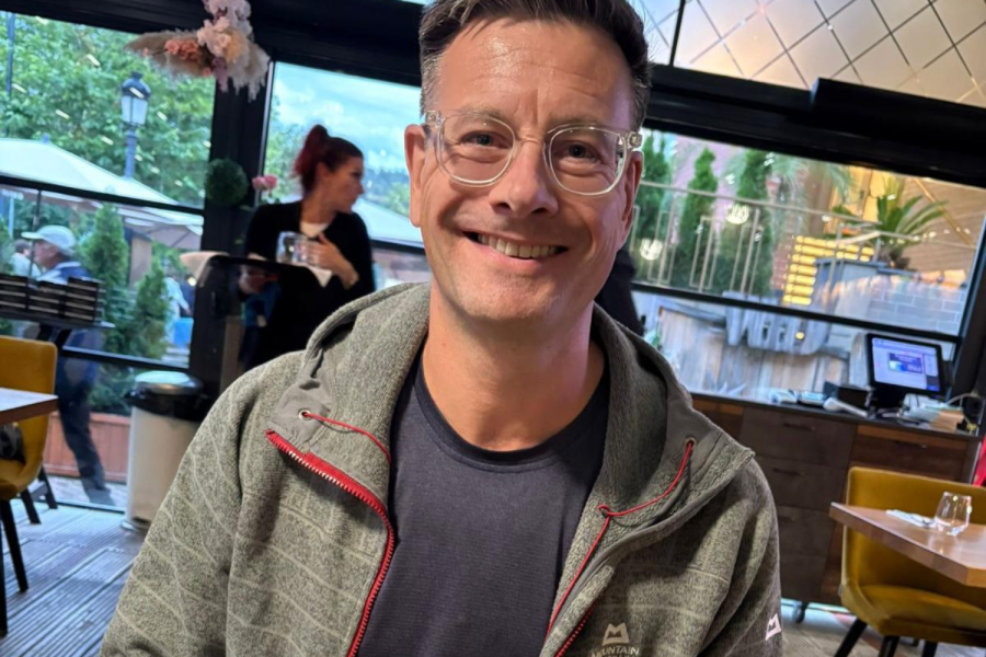 Yr Athro James Hardy yn gwenu ar y camera yn eistedd wrth fwrdd mewn bwyty gyda phryd o'i flaen