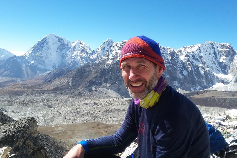 Jonathan Moore yn eistedd ar graig gyda mynyddoedd eiraog y tu ôl iddo, mewn dillad dringo gaeaf cynnes