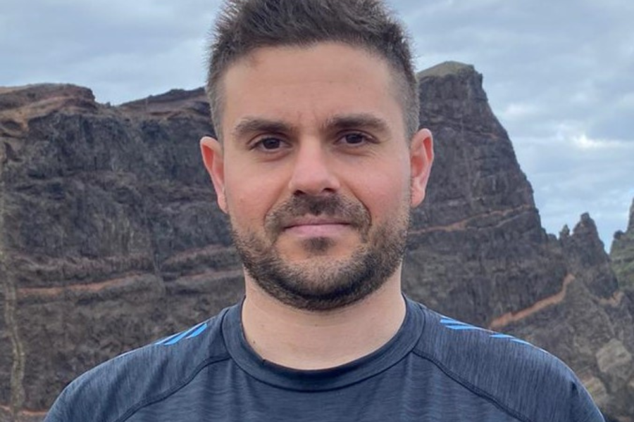 Llun pen a'r ysgwyddau o Germano, mewn crys-t Prifysgol Bangor, o flaen carreg fawr.