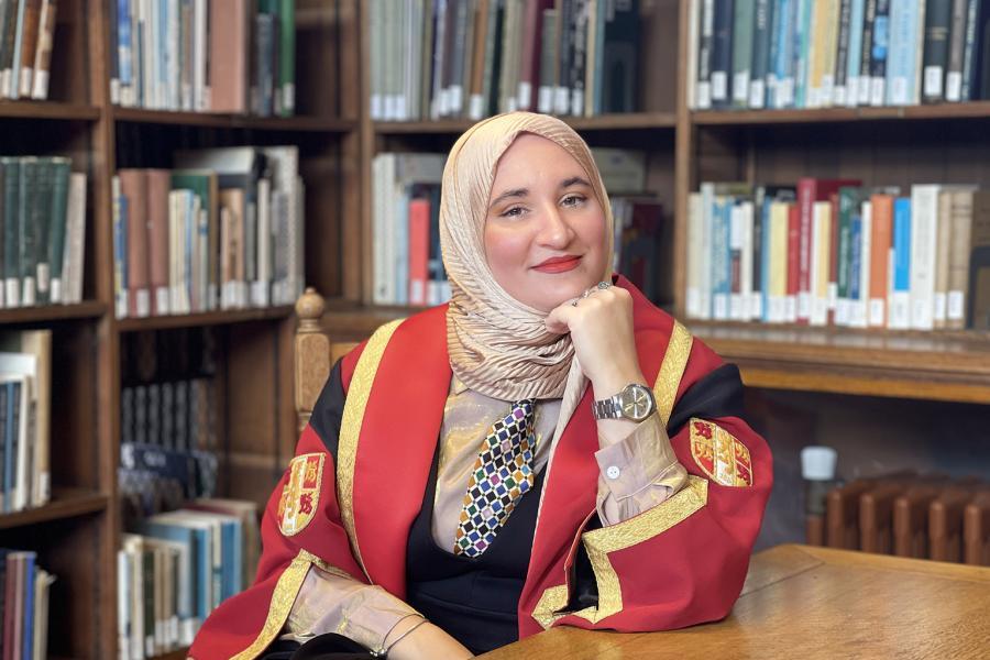 An image of poet Hanan Issa wearing a light-coloured headscarf and a red and gold Bangor University graduation gown sits at a wooden table in a library. They rest one hand against their cheek and wear a patterned tie and a wristwatch. Behind them are tall bookcases filled with neatly arranged books.