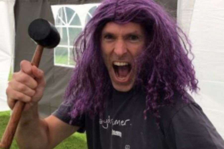 Tim Woodman yn gwisgo wig borffor, yn gwneud wyneb at y camera, yn dal morthwyl