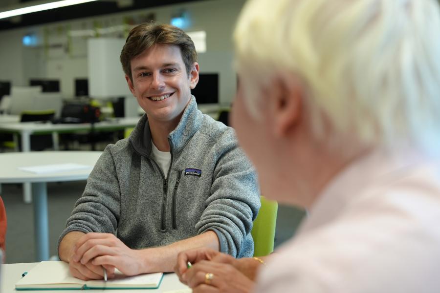A student sits at a desk talking with a lecturer who is sat on the other side of the desk. 