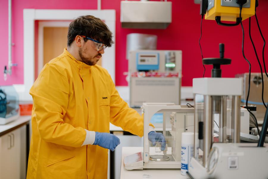 Myfyriwr yn Gweithio yn y Labordy, gyda chôt labordy felen yn mesur bicer