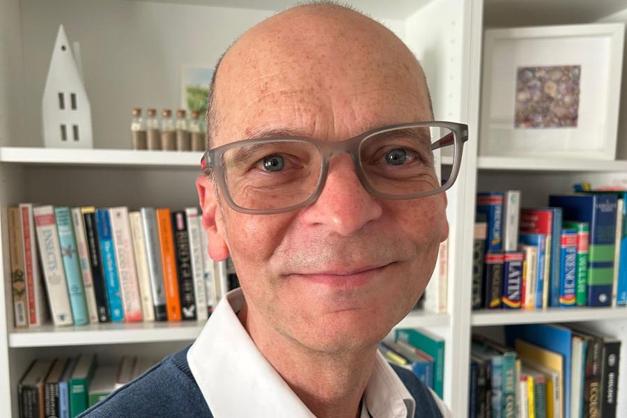 Person yn gwisgo sbectol a chrys â cholêr, yn sefyll dan do o flaen silffoedd llyfrau gwynion llawn llyfrau; mae’r wyneb wedi’i guddio’n fwriadol.