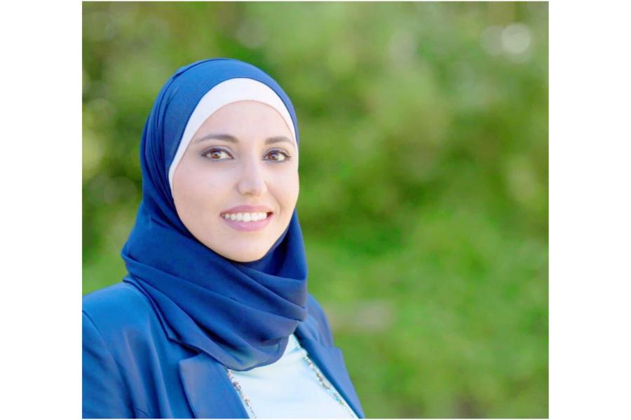 Person wearing a blue headscarf and blue jacket, photographed outdoors against a blurred green background