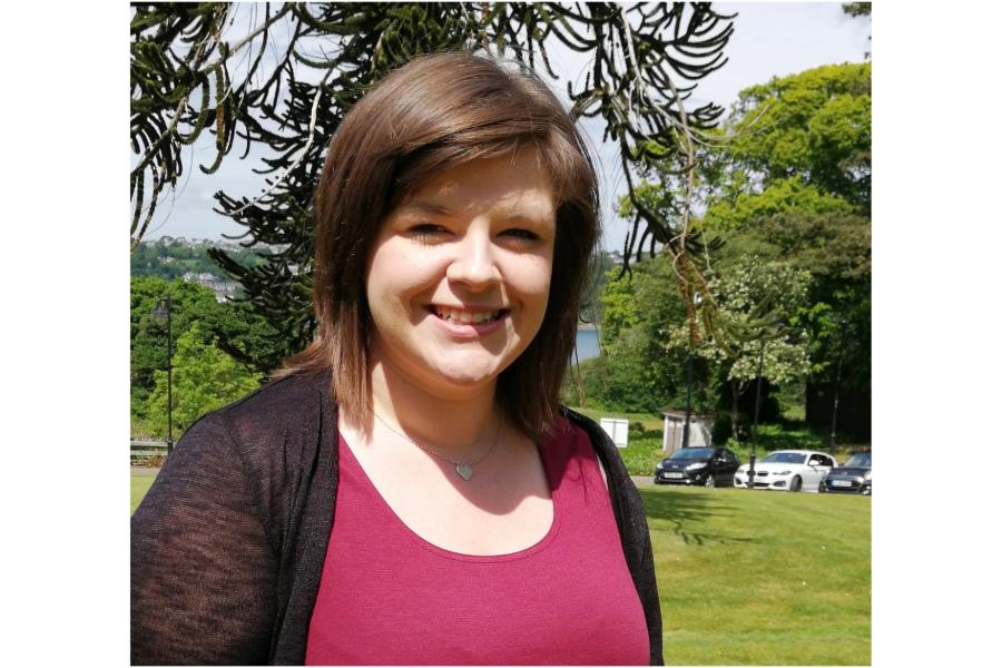 Person standing on a grassy lawn in a park, wearing a sleeveless red top and dark cardigan, with trees and parked cars in the background