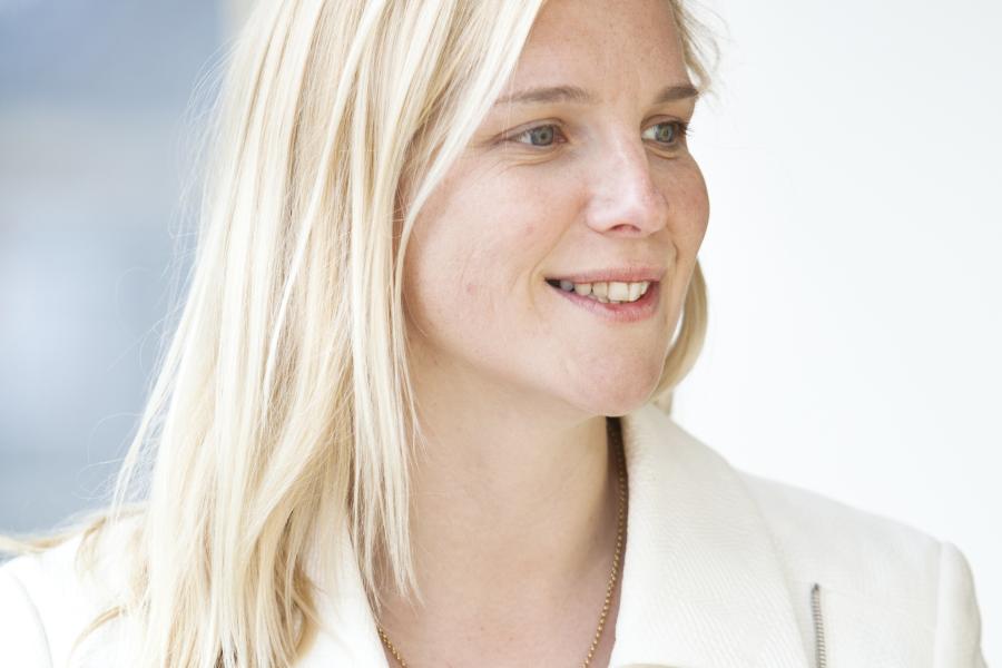 Portrait of a person with shoulder-length blonde hair wearing a light cream jacket, facing slightly to the side against a soft, neutral indoor background.