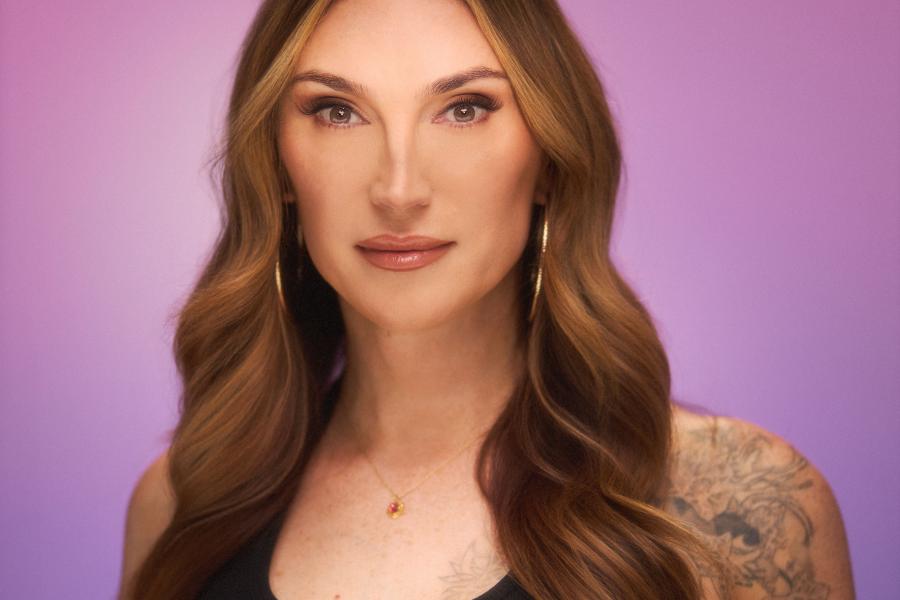 Studio portrait of a person with long wavy brown hair, wearing a black sleeveless top, hoop earrings, and a small pendant necklace, with a tattoo visible on one shoulder, against a soft purple gradient background.