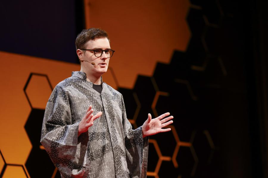 Person giving a talk on a stage, wearing glasses and a patterned loose garment, speaking with both hands raised; orange-lit backdrop with hexagonal shapes.