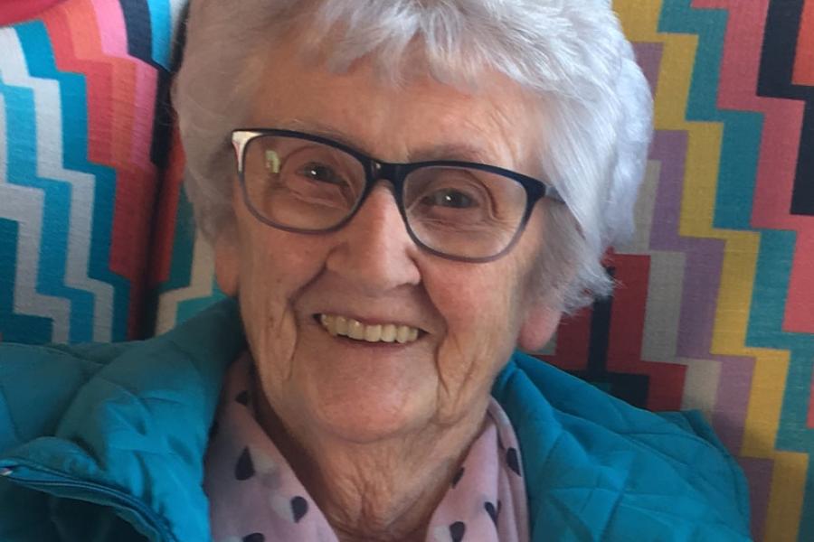 Older adult with short white hair and glasses, seated indoors against colourful patterned chairs, wearing a teal jacket and pink scarf with small droplet shapes.