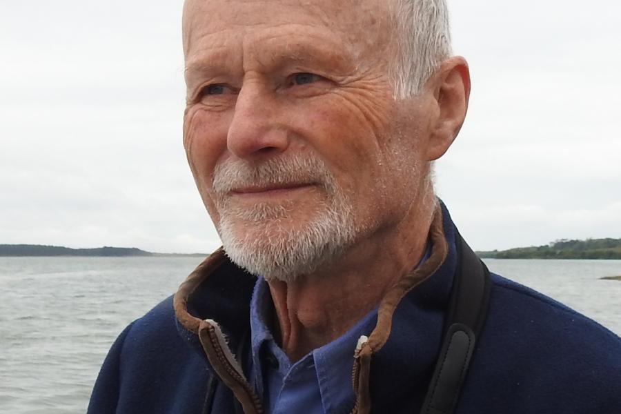 Older adult with short light hair and a trimmed beard, standing outdoors near a calm body of water, wearing a blue jacket over a collared top, with a cloudy sky and distant shoreline in the background.