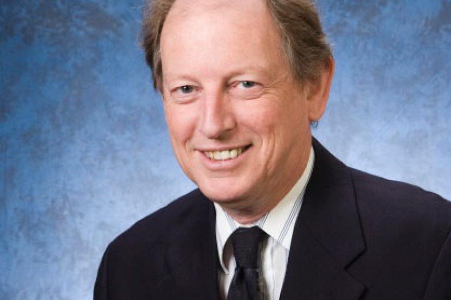 Formal head-and-shoulders portrait of a person wearing a dark suit and tie, facing slightly left, with short hair and a blue studio backdrop.