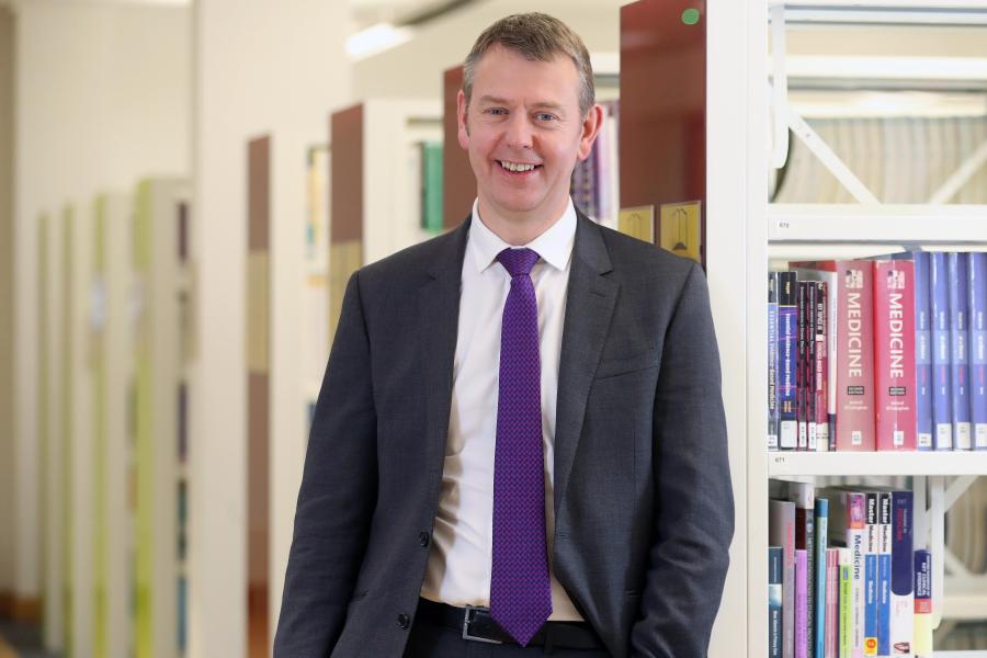 Person in a suit and tie standing in a library aisle beside bookshelves filled with textbooks, facing the camera.
