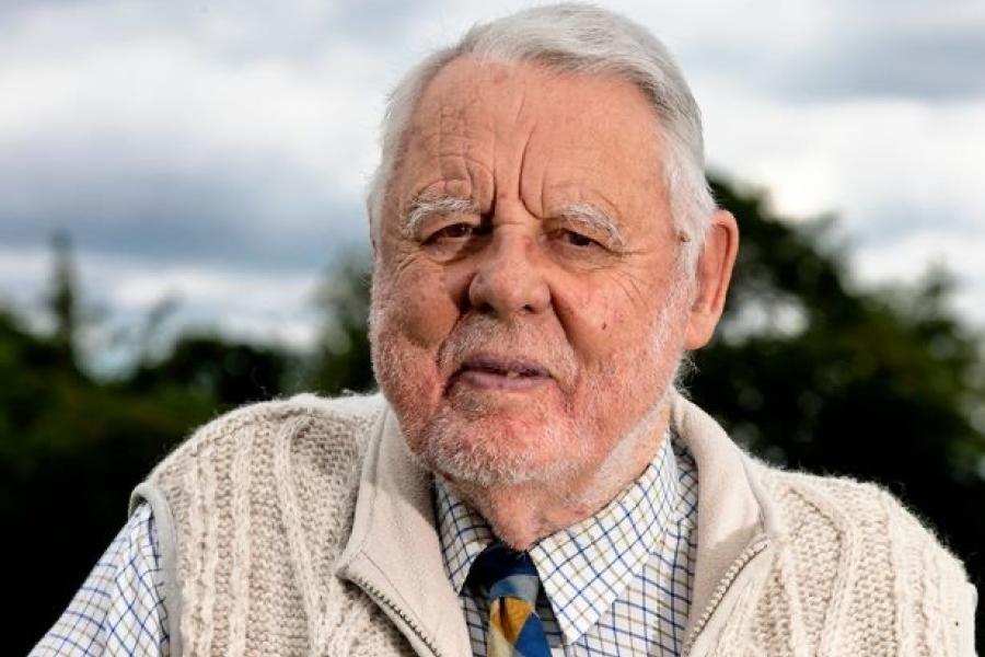 Elderly person outdoors, wearing a cream cable-knit cardigan over a checked shirt and striped tie, facing the camera with trees and cloudy sky in the background.