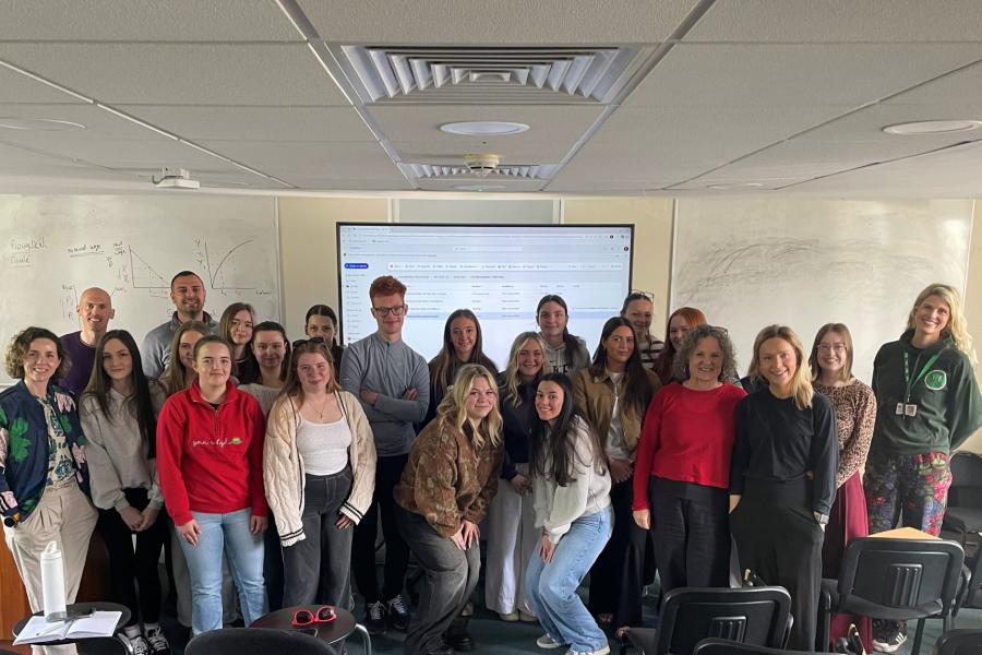 Myfyrwyr a staff wedi ymgynnull mewn ystafell ddosbarth, yn sefyll ac yn cwrcwd mewn grŵp o flaen bwrdd gwyn a sgrin wedi’i gosod ar y wal sy’n dangos tudalen we, gyda rhesi o gadeiriau gwag yn y blaendir a goleuadau a fentiau ar y nenfwd uwchben.