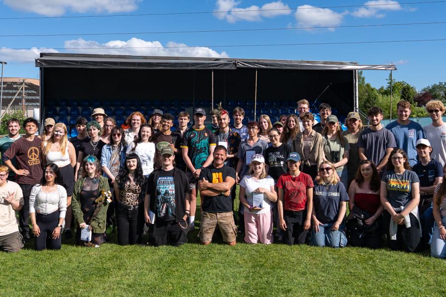 A large group posed in several rows on grass in front of a temporary stage and seating area, with trees and a clear blue sky in the background.