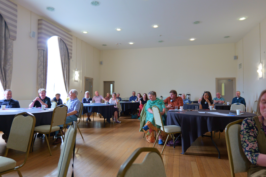 Cyfranogwyr yn eistedd o amgylch byrddau mewn neuadd ddigwyddiadau fawr yn ystod digwyddiad dathlu cyrsiau byr Prifysgol Bangor. Mae'r mynychwyr yn gwrando ac yn cymdeithasu mewn ystafell olau ac eang.