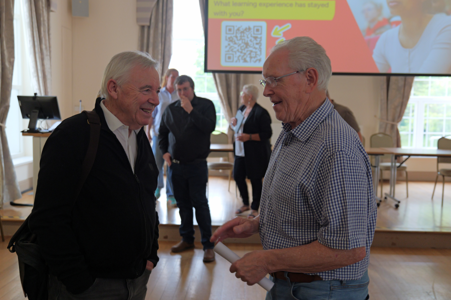 Dau fynychwr yn sgwrsio yn y neuadd ddigwyddiadau yn ystod digwyddiad dathlu cyrsiau byr Prifysgol Bangor. Mae cyfranogwyr eraill i’w gweld yn y cefndir ger llwyfan a sgrin gyflwyno sy’n dangos sleid am brofiadau dysgu.