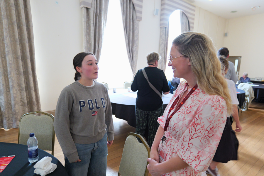 Dau fynychwr yn sefyll ac yn sgwrsio mewn neuadd ddigwyddiadau olau yn ystod digwyddiad dathlu cyrsiau byr Prifysgol Bangor, gyda chyfranogwyr eraill yn cymdeithasu ac yn trafod yn y cefndir. Mae byrddau, cadeiriau a deunyddiau’r digwyddiad i’w gweld o amgylch yr ystafell.