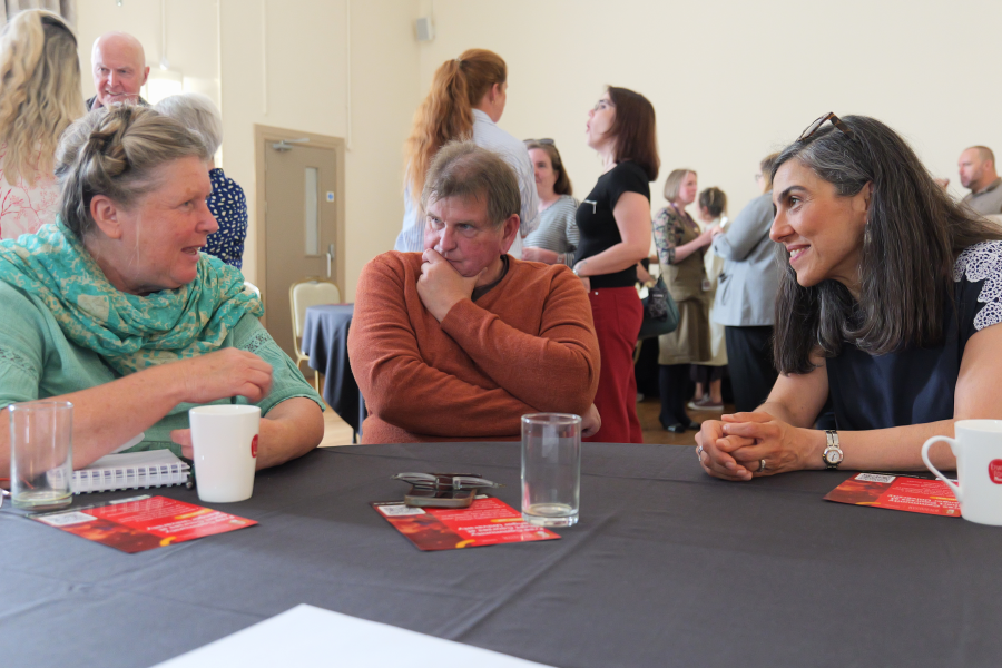 Sawl mynychwr yn eistedd o amgylch bwrdd ac yn cymryd rhan mewn sgwrs yn ystod digwyddiad dathlu cyrsiau byr Prifysgol Bangor. Mae cyfranogwyr eraill yn sefyll ac yn rhwydweithio yn y cefndir, gyda thaflenni gwybodaeth a lluniaeth i’w gweld ar y bwrdd.