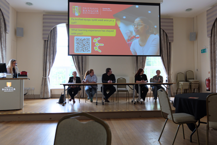 Panel o gyfranogwyr cyrsiau yn eistedd ar flaen neuadd ddigwyddiadau tra bo aelodau o'r gynulleidfa yn gwrando. Mae sgrin fawr y tu ôl i'r panel yn dangos sleid gyflwyno gyda brandio Prifysgol Bangor a chwestiynau am brofiadau'r dysgwyr.