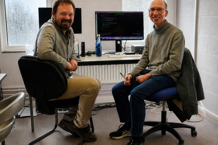Two men sitting on chairs next to a desk with two computers on it