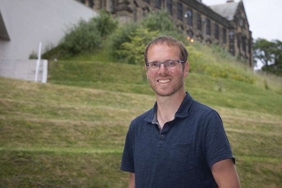 Dyn efo spectol yn gwisgo crys polo glas, mae gwair a Prif Adeilad Prifysgol Bangor yn y cefndirsmiles into camera- with a background of cut grass and Bangor University building in the background.