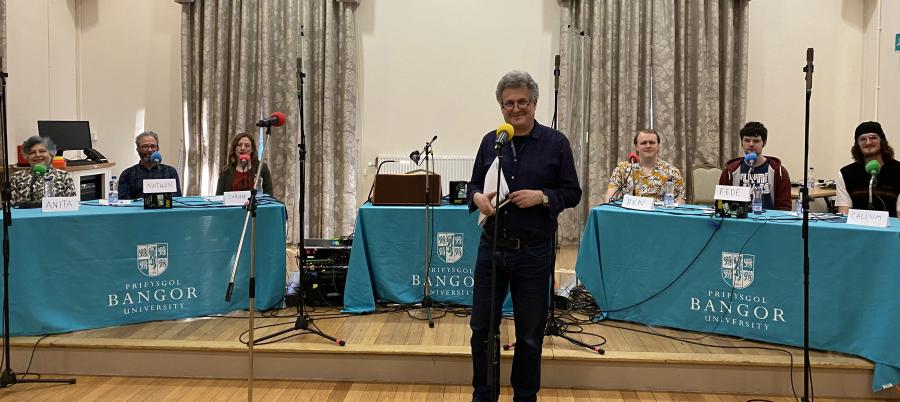 Bangor University 3rd degree Quiz members sit behind tables and microphones ready to compete. In the front is producer, David Tyler.