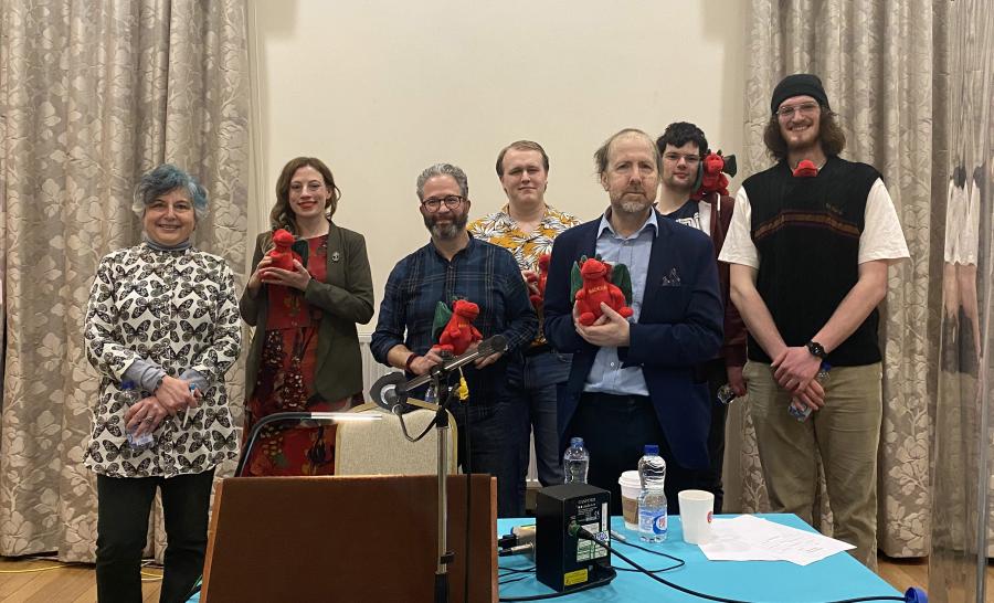 A group of people standing behind a table and microphone, they hold small red dragon soft toys