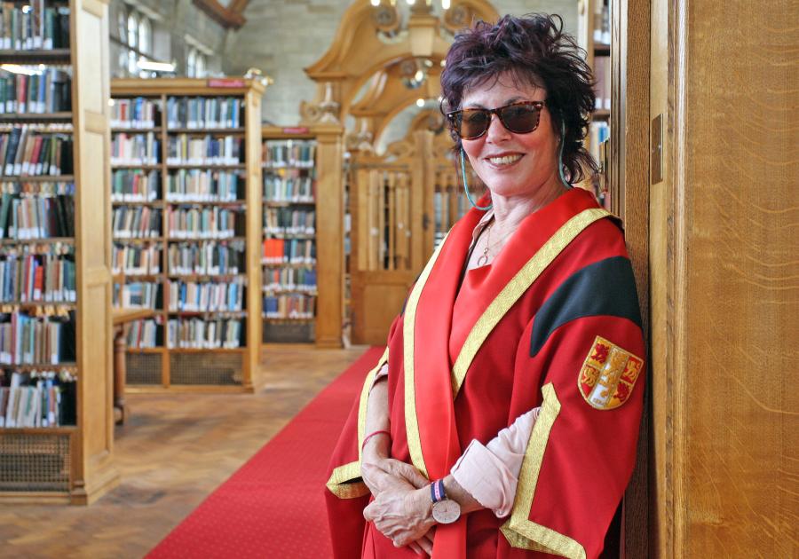 Ruby wax in red academic gown and dark glasses, leans on the end of a book shelf in the Shankland Library at Bangor University