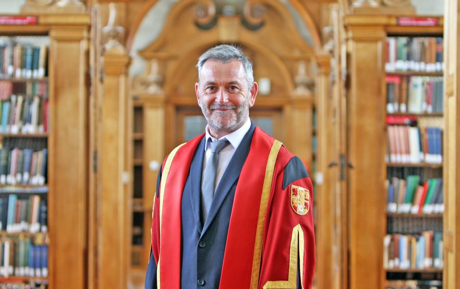 Besuited man stands in red academic gown in Library