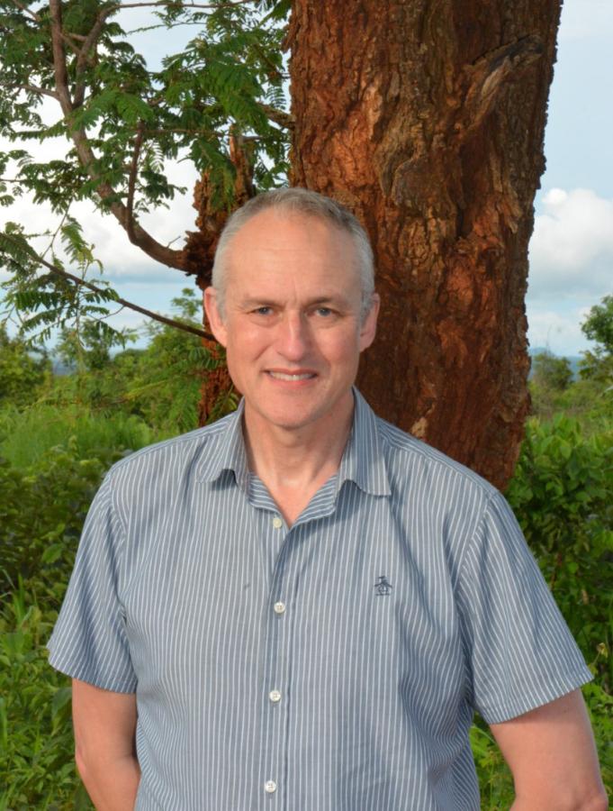 person in a short sleeved shirt standing in an outdoor landscape with a tree and shrubbery in the background