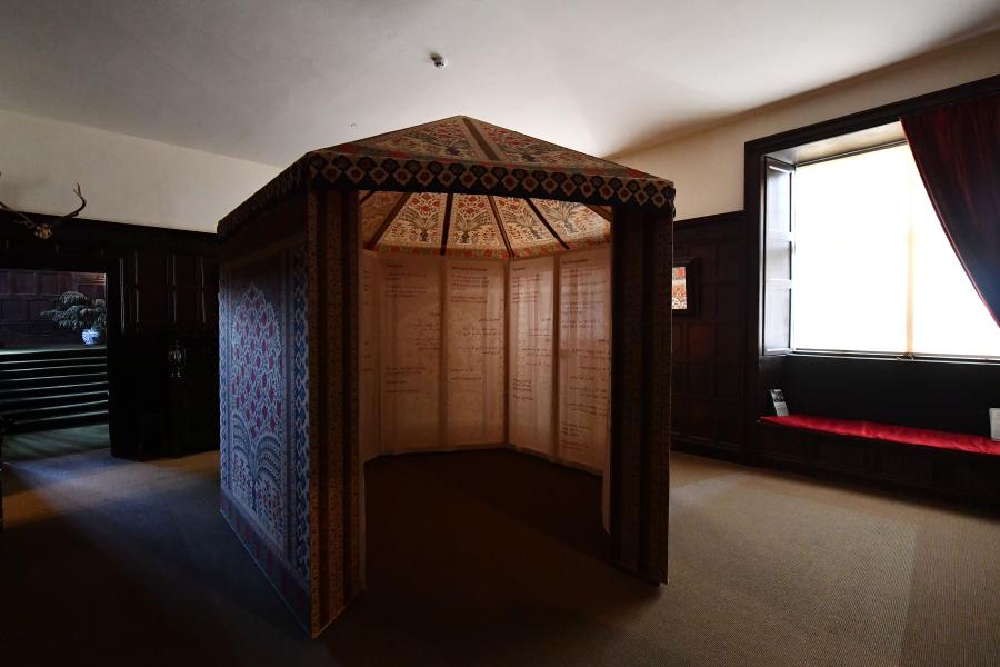 A tent with painted floral decoration on the outside and writing on the inside sits in the centre of a grand room.
