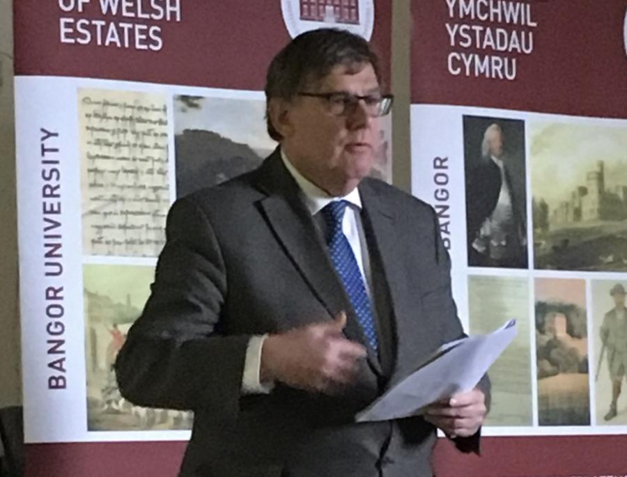 Llun o Gwilym Owen yn cyflwyno, yn sefyll o flaen baner ISWE