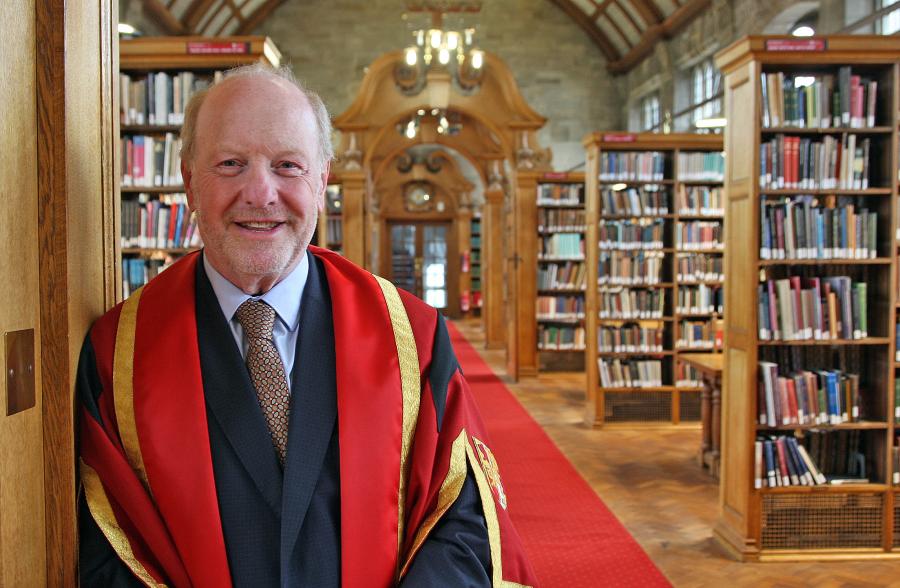 A man wearing red and gold graduation robe leaning on wooden pillar in a library with bookshelves in the background