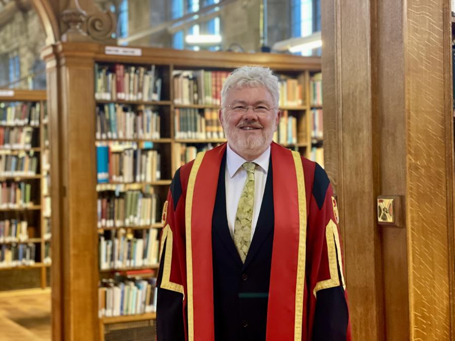Image of Professor David N Thomas wearing red and gold honorary graduate gowns. He is standing in Shankland Reading Room
